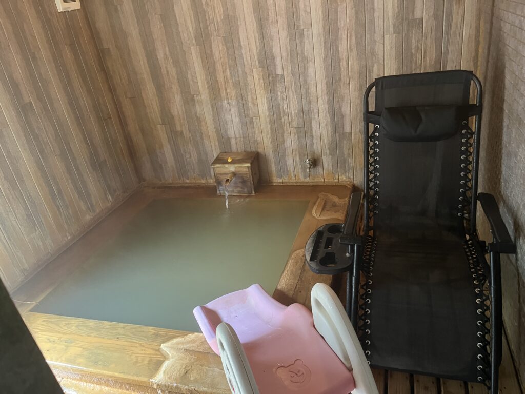 季一湯「九ノ湯」の内湯。源泉が注がれる石造りの浴槽と、その傍らに設置された子供用のピンクの滑り台、そして大人が休憩できるリクライニングチェアが並ぶ家族風呂の様子。